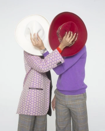 Two people stand closely together, obscuring their faces with a large white and red hat, wearing blazers and patterned pants on a white backdrop.