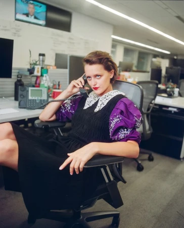 Stylish woman in office chair answers phone with television and office equipment behind. Purple, black, and white dress.