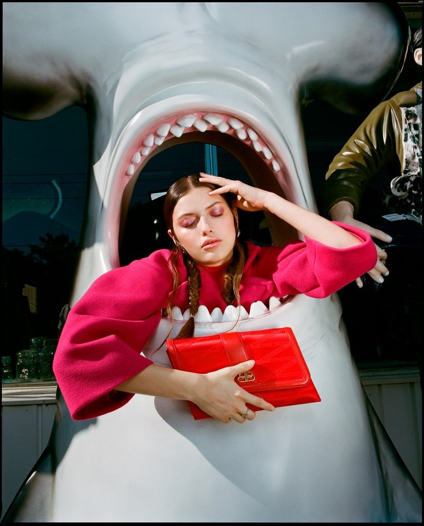 A young woman in a pink top is framed by the mouth of a large shark sculpture, holding a red clutch bag in a surreal fashion portrait.