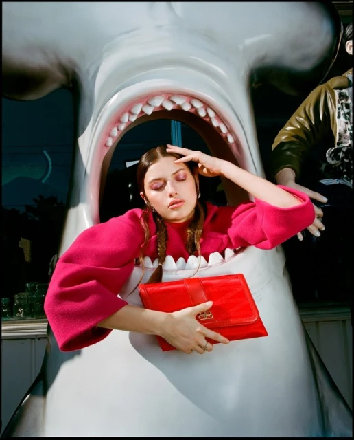 A young woman in a pink top is framed by the mouth of a large shark sculpture, holding a red clutch bag in a surreal fashion portrait.