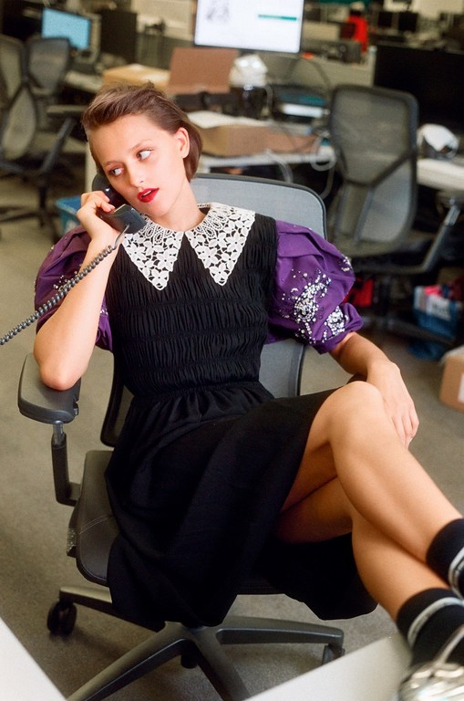 Young woman in a black dress with a white collar and purple sleeves talking on the phone in a modern, bright, open-plan office space.