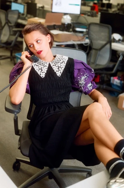 Young woman in a black dress with a white collar and purple sleeves talking on the phone in a modern, bright, open-plan office space.