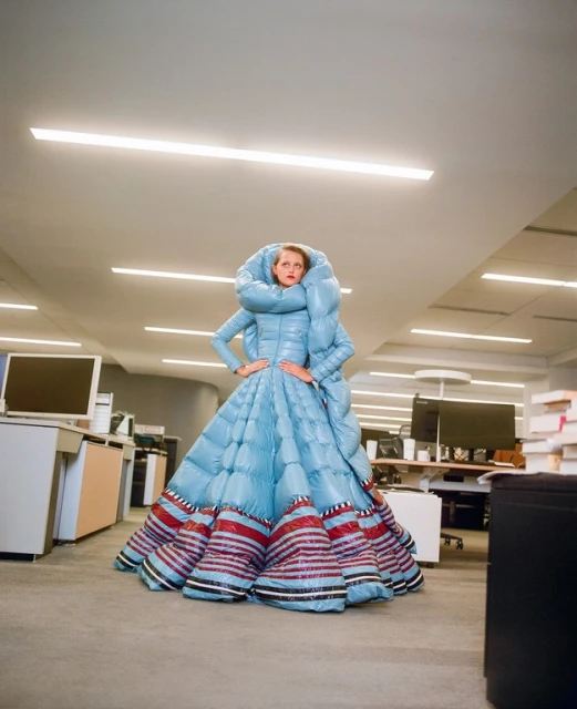 Fashionable adult female poses in a large, light-blue quilted puffer dress with colorful hem stripes inside a modern office setting.