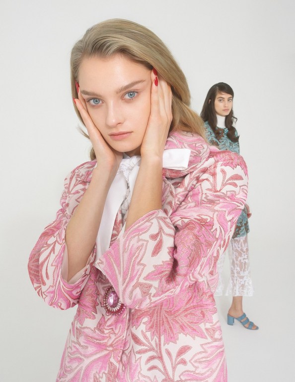 Two women in studio, one with blonde hair in the foreground wearing a pink floral coat and the other in background.