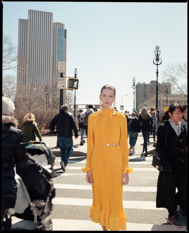A woman in a bright yellow dress walks across a crosswalk in an urban setting with a high-rise building and pedestrians nearby.
