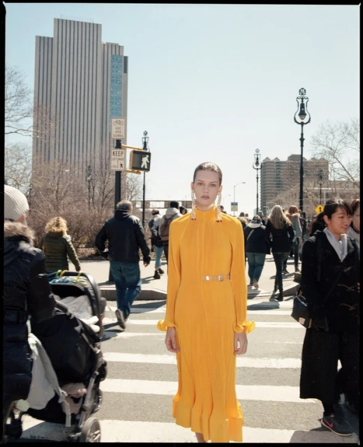 A woman in a bright yellow dress walks across a crosswalk in an urban setting with a high-rise building and pedestrians nearby.