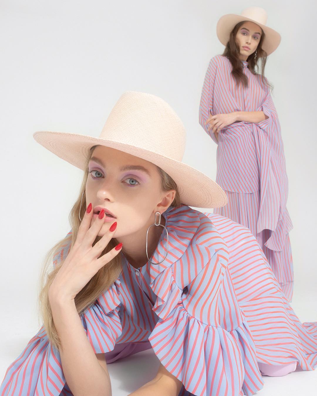 Two fair-skinned young women in matching pink-and-blue striped dresses and straw hats pose in a brightly lit photo studio setting.