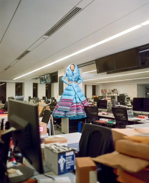Woman posing in avant-garde blue puffer gown in office. The open space has computers, furniture, and general office clutter. 