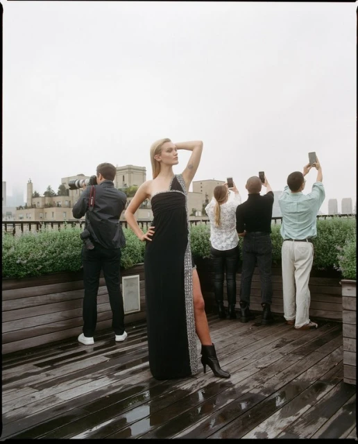 Fashion shoot on a rooftop: a blonde model in a black dress poses for photographers against a city skyline on an overcast day.