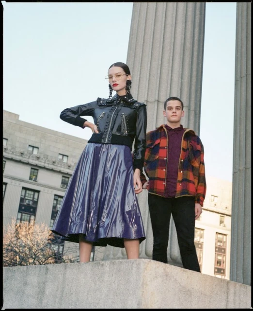 Two people stand next to a concrete column outdoors. An adult female in a black jacket and purple skirt poses with a young adult male.
