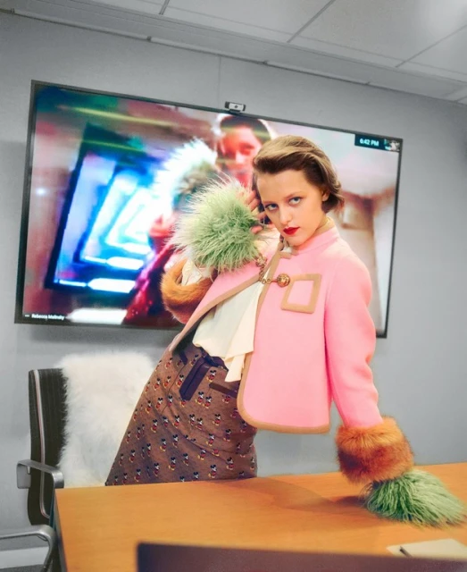 Stylish young woman poses in a pink jacket with fur cuffs and trim in front of an office conference room with an active video call.