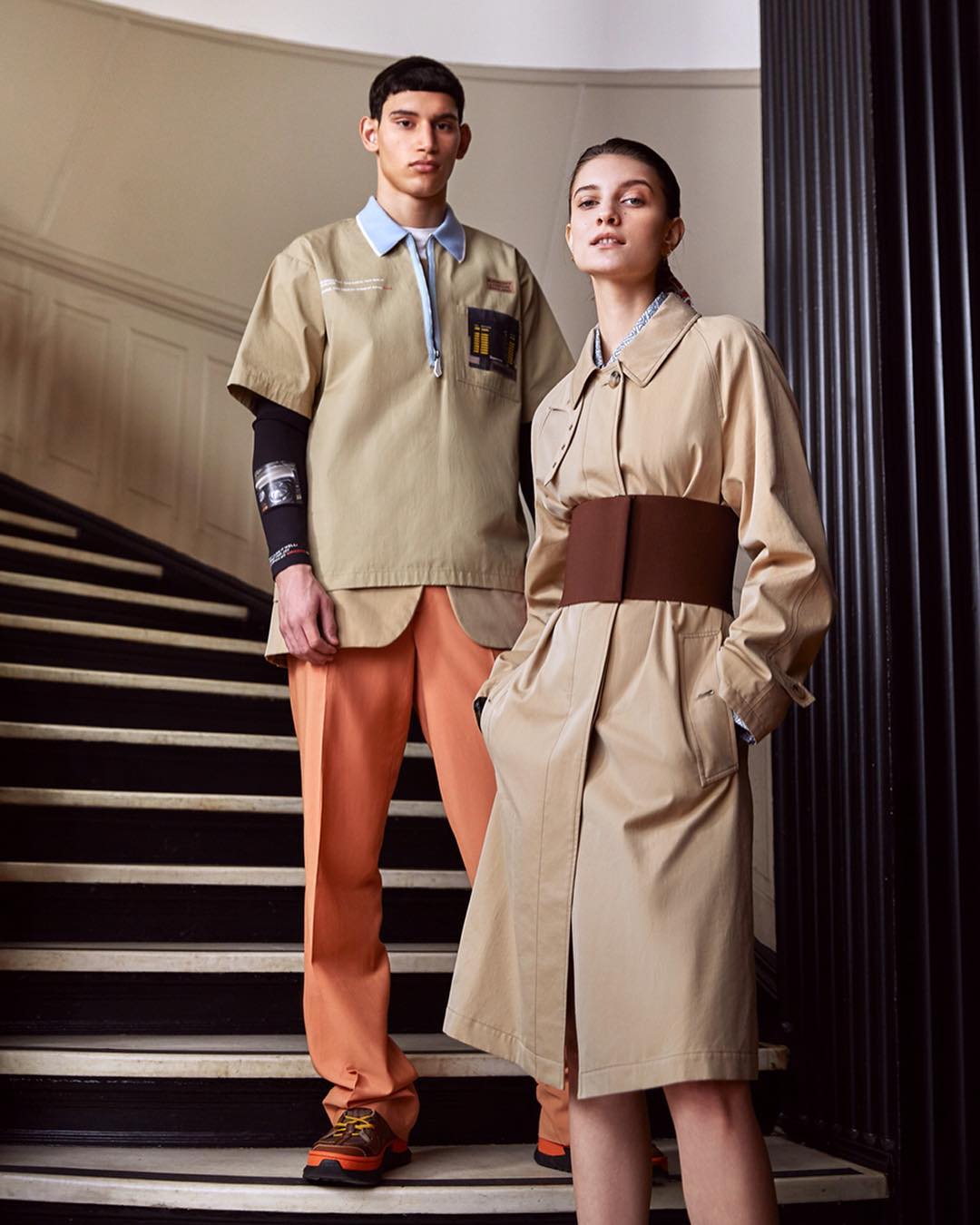 Two fashion models pose on stairs indoors. The man wears a khaki shirt, orange pants, and hiking shoes. The woman wears a beige trench coat and wide brown belt.