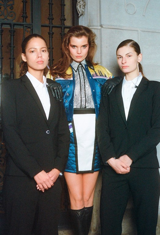 Three women stand in a fashion-editorial style image. One woman in a colorfully patterned jacket stands between two women in dark suits.
