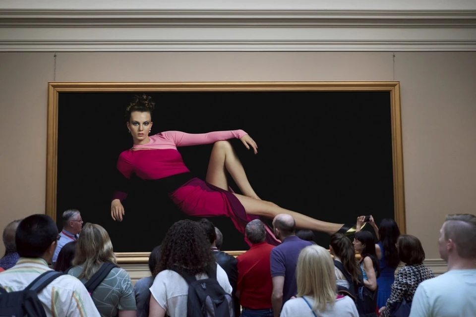 Museum visitors view a large portrait of a woman in a pink and red dress against a black background, inside an art gallery with beige walls.