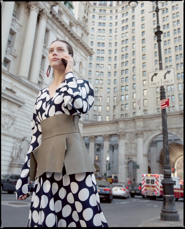 Woman in polka dot dress uses her cell phone in an urban environment. Buildings, traffic and ambulance are visible in the background.