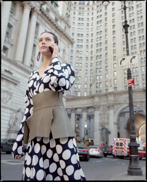 Woman in polka dot dress uses her cell phone in an urban environment. Buildings, traffic and ambulance are visible in the background.
