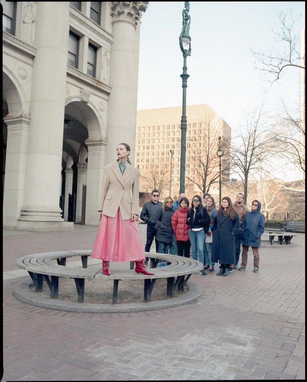 Fashion shot in city square: a model in a tan blazer and pink skirt stands on a bench, surrounded by a group of onlookers.