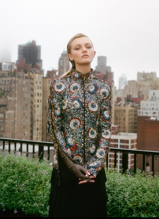 Woman in patterned jacket poses on rooftop with city background. Neutral expression, outdoor fashion shot, overcast day, natural light.