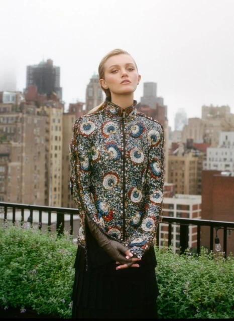 Woman in patterned jacket poses on rooftop with city background. Neutral expression, outdoor fashion shot, overcast day, natural light.