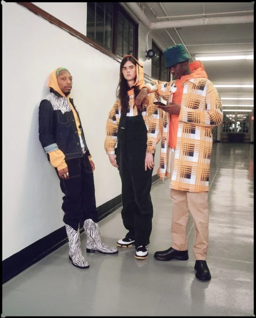 Three people in patterned jackets and streetwear fashion stand indoors in an hallway, photographed at eye-level in medium shot.