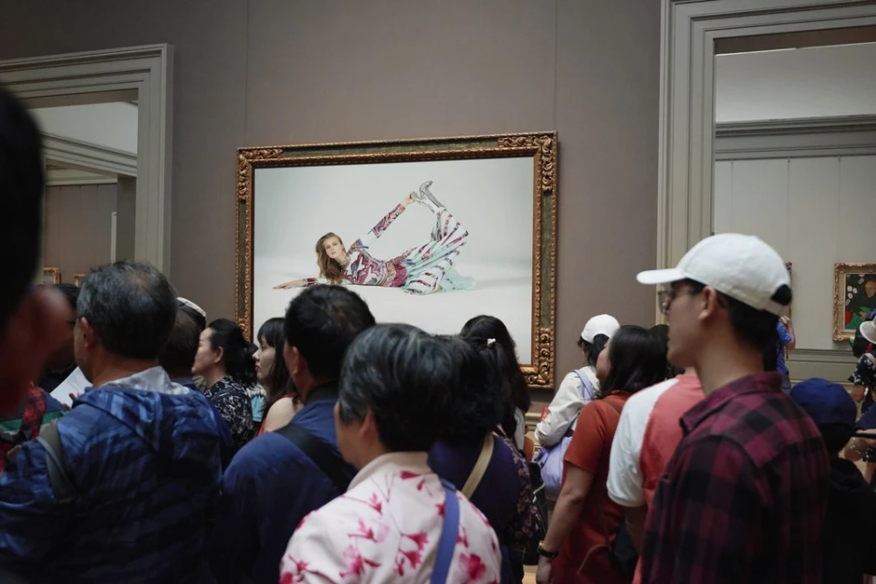 Museum visitors view a large framed photograph of a model in a patterned jumpsuit reclining against a white backdrop on a gallery wall.