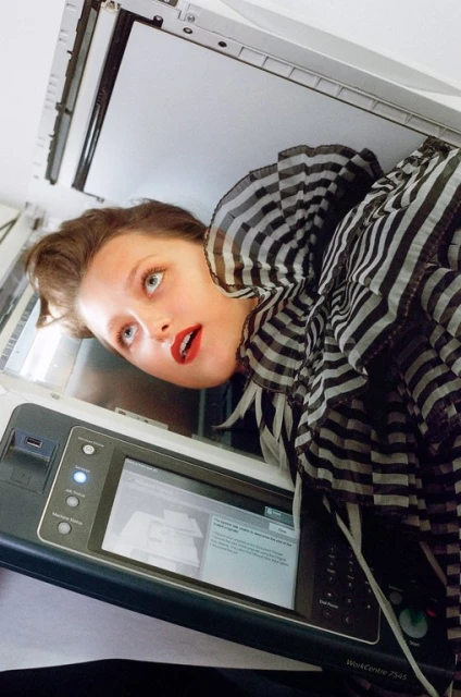 Woman peers out from a digital printer in an indoor setting, wearing a striped scarf and red lipstick, showcasing a creative, unconventional composition.