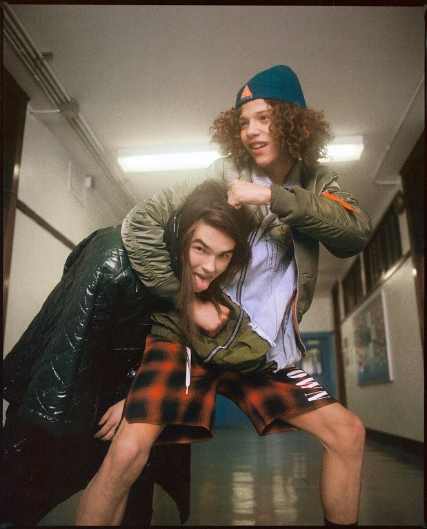 Two young men playfully wrestling in an indoor hallway with overhead fluorescent lighting and bright walls, showcasing casual fashion styles.