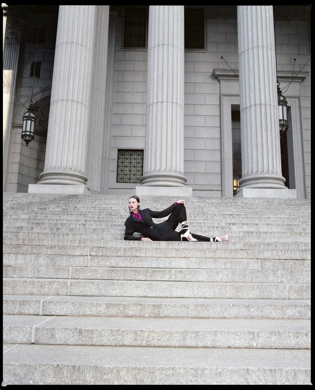 Woman in a black suit and heels lounges on the gray stone steps of a neoclassical building with tall, fluted Doric columns.