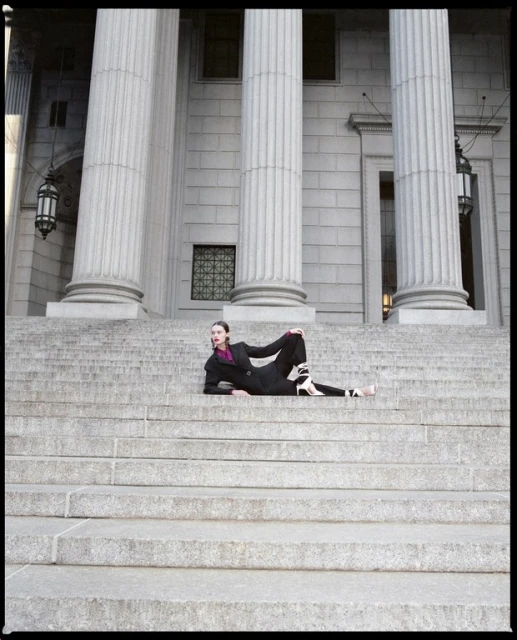Woman in a black suit and heels lounges on the gray stone steps of a neoclassical building with tall, fluted Doric columns.