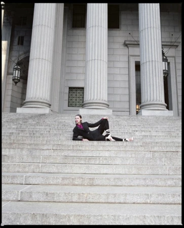 Woman in a black suit and heels lounges on the gray stone steps of a neoclassical building with tall, fluted Doric columns.