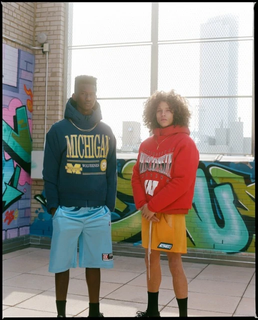 Two young men pose in collegiate apparel on a rooftop with graffiti, featuring Michigan and Wisconsin designs, shorts, and a city view.