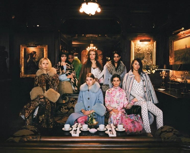 Group of eight women posing indoors in a vintage-style room wearing winter coats and dresses in front of table setting with tea cups.