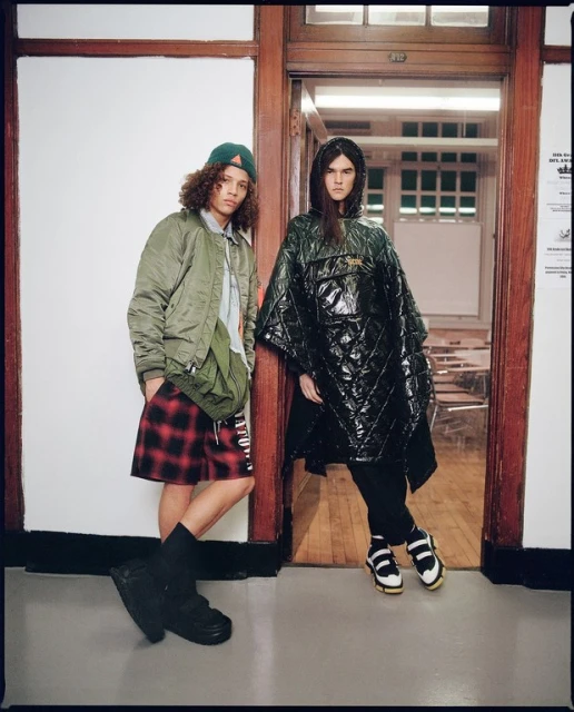 Two models pose indoors; one wears an olive jacket and plaid shorts, the other a quilted poncho and sandals, fashion-forward and stylish.
