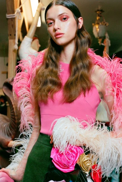 Headshot of a female fashion model with long brown hair, pink top and statement feather boa around her shoulders and waist.