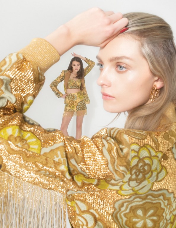 Two women are captured in a studio portrait. One woman is up close wearing a gold jacket. Another woman wears a gold matching set.