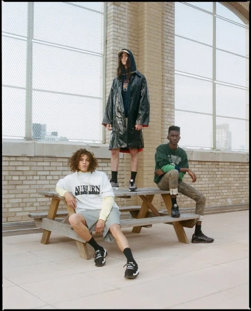 Three adult fashion models wear trendy casual streetwear outfits and pose on a rooftop in front of a city skyline and tall windows.