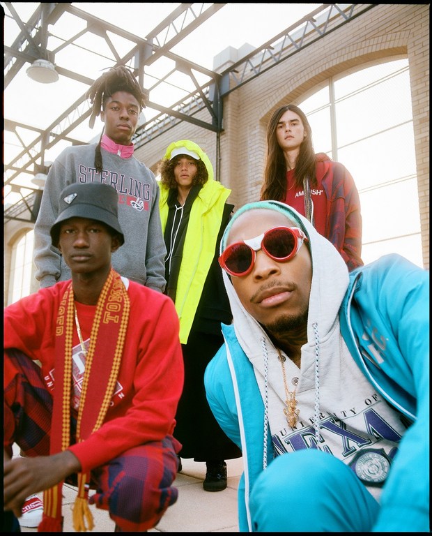 A group of five fashionably dressed young adults pose in an open industrial building, showcasing streetwear with bold colors and unique accessories.