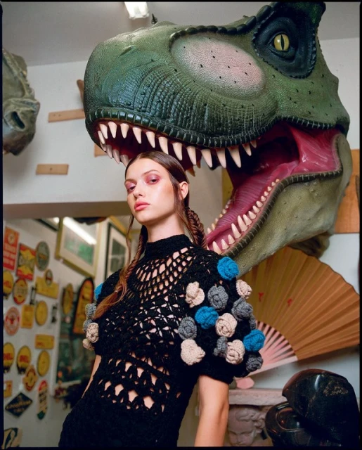 Young woman posing in a black crochet dress with decorative pom poms. A large dinosaur head sculpture sits behind her indoors.