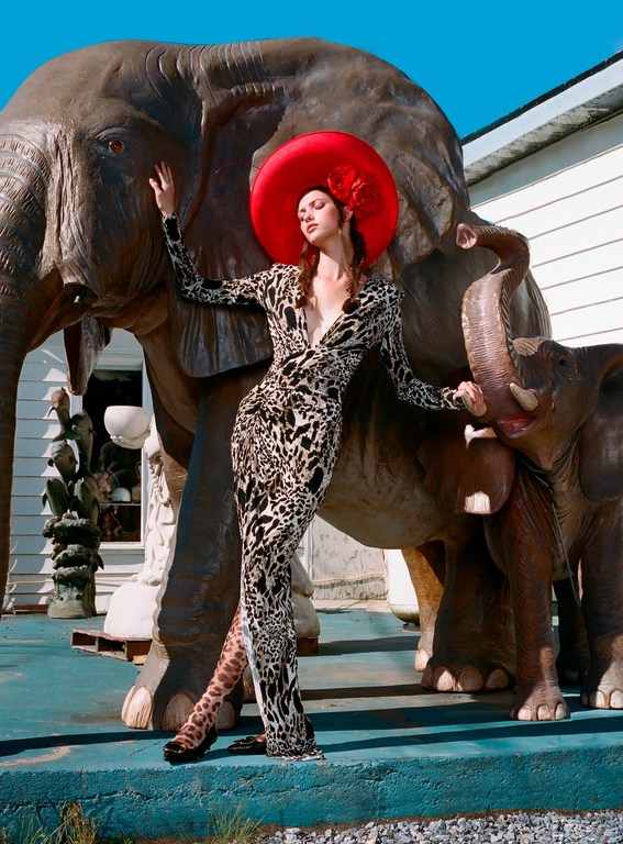 A woman in a patterned dress and a large red hat poses next to elephant statues outside, illuminated by bright daylight.