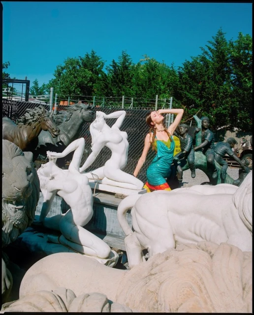 A woman in a green dress stands among an assortment of white and bronze sculptures of horses, figures, and a lion, set against a blue sky.
