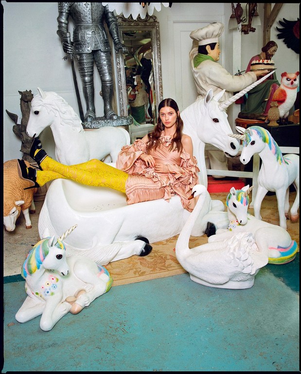 Woman in a pink dress reclines among white unicorn and swan figurines inside an ornate room with a vintage mirror and decor.