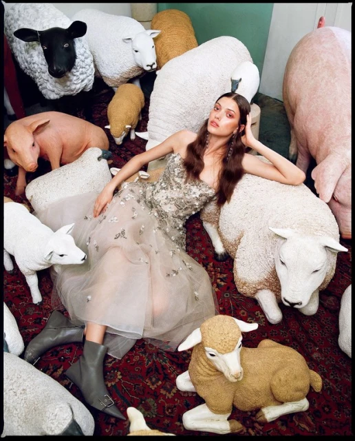Woman in embellished gown surrounded by faux sheep, one black sheep, on red patterned rug in studio. Fashion editorial photo shoot.