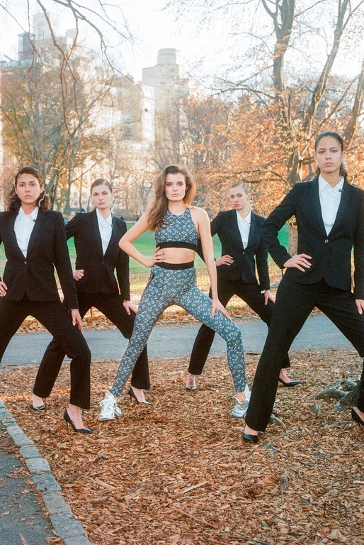 Five women pose outdoors on a path surrounded by trees and a building; one wears activewear, four wear black business suits.
