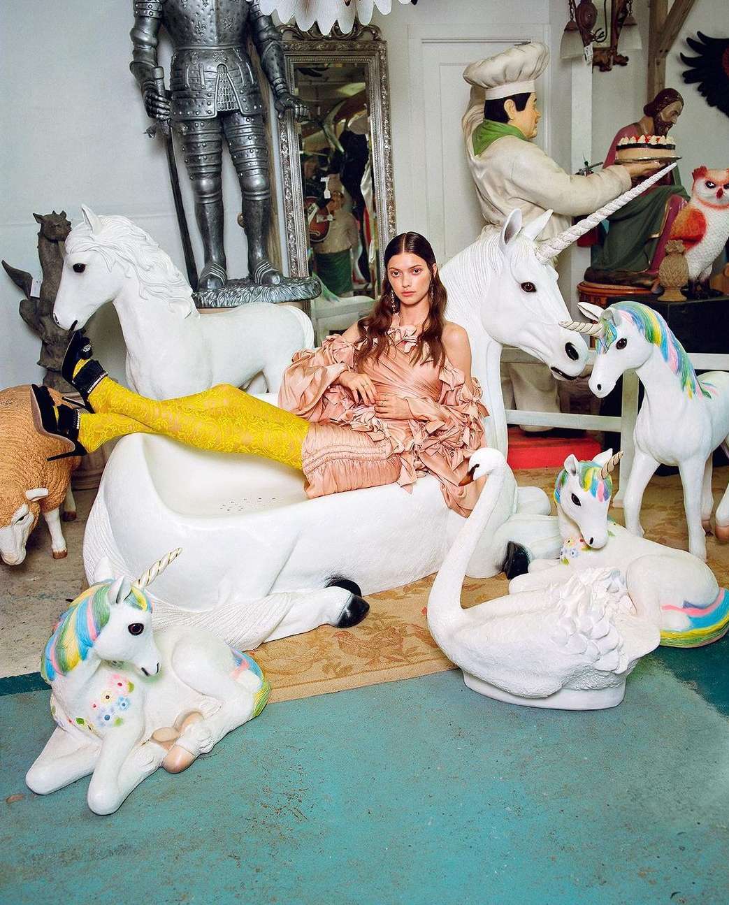 Eye-level shot of woman in dress and yellow tights reclining among white unicorn and swan figurines in an indoor setting.