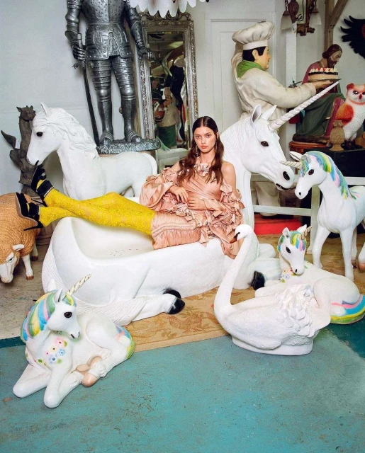 Eye-level shot of woman in dress and yellow tights reclining among white unicorn and swan figurines in an indoor setting.