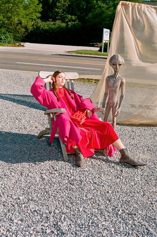 A woman in a vibrant pink outfit lounges in a chair next to a grey alien figure in an outdoor setting with a road and trees.