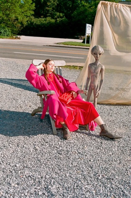 A woman in a vibrant pink outfit lounges in a chair next to a grey alien figure in an outdoor setting with a road and trees.