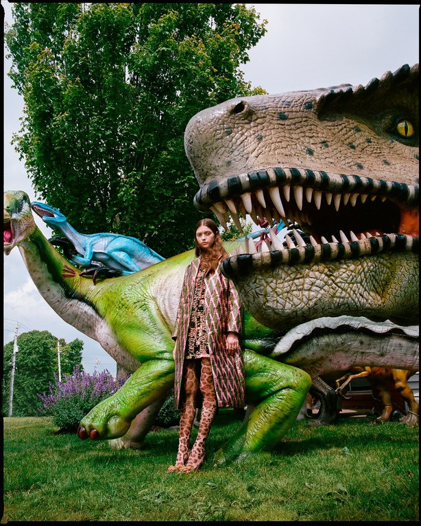Adult female poses in printed coat and tights, with decorative dinosaur statues, outdoors on a green grass lawn, overcast sky.