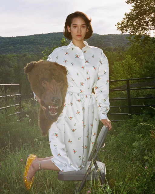 Woman in floral dress sits on a chair outdoors holding a bear head mask, set against a backdrop of grassy fields and distant hills.