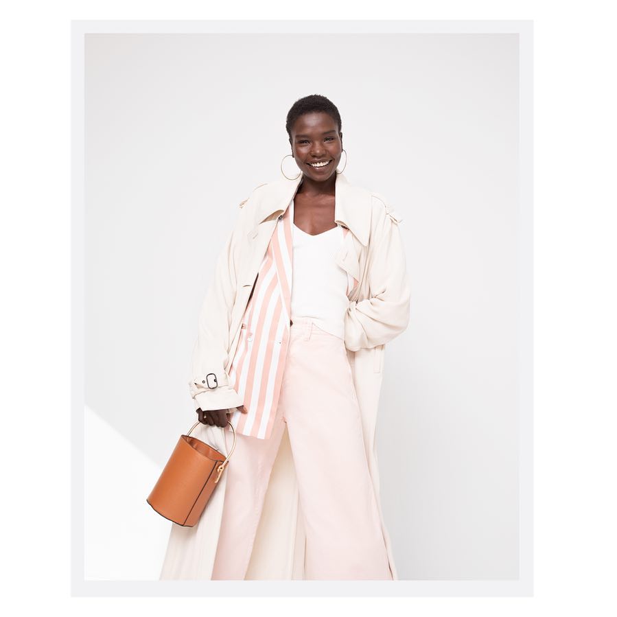 Smiling Black woman in a cream trench coat, striped blazer, and pink pants holding a brown bucket bag against a white background.